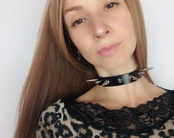 May include: Black leather choker necklace with silver spikes. The choker is worn by a person with long brown hair and a leopard print shirt.