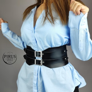 Puede incluir: Un cintur&oacute;n de cuero negro de doble hebilla con hebillas plateadas. El cintur&oacute;n se lleva sobre una camisa de botones azul claro.