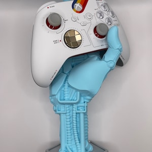 May include: A white Xbox controller with a rainbow design is held by a blue 3D printed robot hand. The robot hand is attached to a blue 3D printed base.
