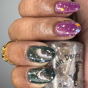 May include: A clear glass bottle of nail polish with a white label that says "Rain Dance" and a silhouette of a person holding an umbrella. The bottle is 15 ml. The bottle is next to a hand with green and purple nail polish with glitter.