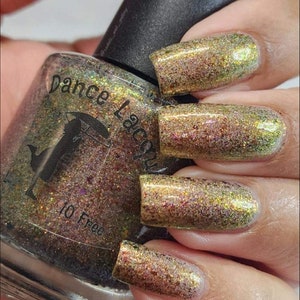 May include: A clear glass bottle of nail polish with a black cap. The bottle is labeled "Dance Lacquer" and "10 Free". The polish is a gold color with a shimmery finish. The bottle is shown with a hand wearing the polish on all five fingernails.