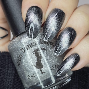 May include: A clear glass bottle of silver glitter nail polish with a black cap. The bottle label reads "Rain Dance Lacquer" and "15 ml". The bottle is shown with a hand wearing silver glitter nail polish on all five fingernails.