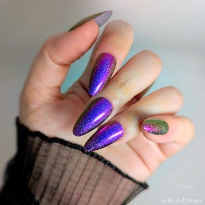 May include: Close-up of long, iridescent purple and blue nail polish on long, almond-shaped nails. The thumb nail is a muted gray-green. The image includes the text "nails.and.hamer".