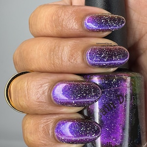 May include: Close-up of a hand with long, oval-shaped nails painted with a shimmering purple nail polish. The polish has a gradient effect, transitioning from dark purple to a lighter shade with silver glitter. A black nail polish bottle is held in the hand.