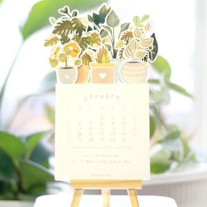 May include: A small desktop calendar with a January 2023 calendar page. The calendar is decorated with a paper cutout of potted plants in shades of green, yellow, and brown.