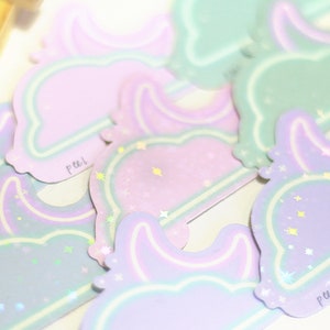 May include: A set of pastel-colored stickers with a moon and star design. The stickers are holographic and have a peel-off backing. The stickers say "peel" on the bottom.