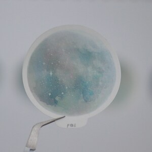 Puede incluir: Una pegatina transparente con un dise&ntilde;o de acuarela de una galaxia azul y verde con estrellas blancas. La pegatina tiene la palabra "pal" escrita en ella.