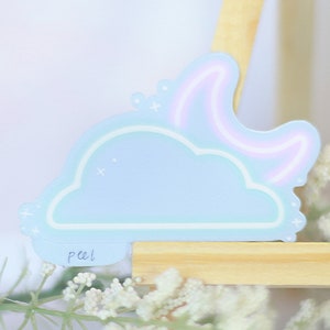 May include: A light blue sticker with a cloud and a crescent moon with a pink neon outline. The sticker has a white outline and a "peel" label on the bottom.