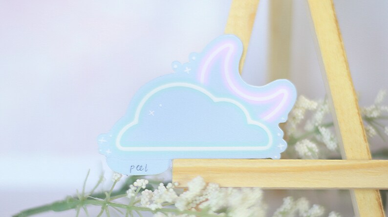 May include: A light blue sticker with a cloud and a crescent moon with a pink neon outline. The sticker has a white outline and a "peel" label on the bottom.