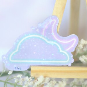 May include: A holographic sticker with a purple cloud and a pink crescent moon. The sticker has a glittery finish and says "peel" in the bottom left corner.