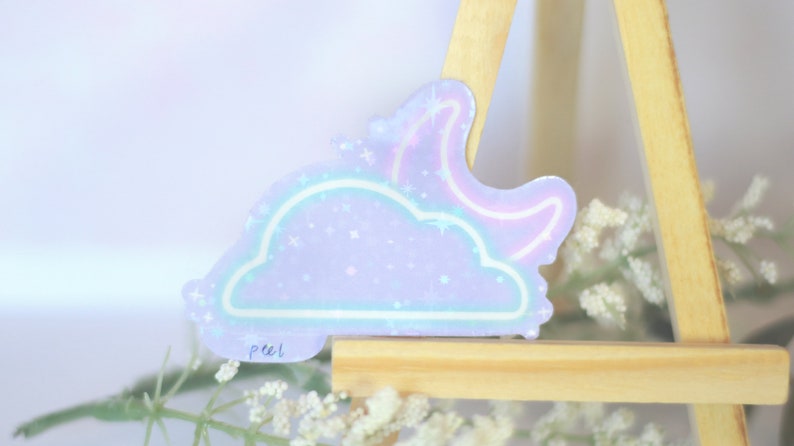May include: A holographic sticker with a purple cloud and a pink crescent moon. The sticker has a glittery finish and says "peel" in the bottom left corner.