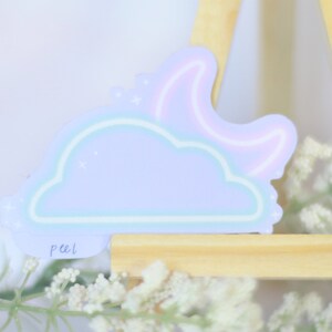 May include: A pastel blue cloud sticker with a pink crescent moon and stars. The sticker has a peel tab and is on a wooden easel.