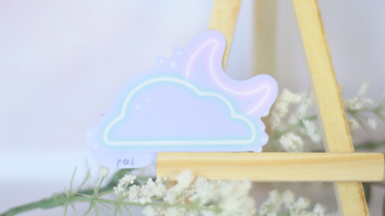May include: A pastel blue cloud sticker with a pink crescent moon and stars. The sticker has a peel tab and is on a wooden easel.