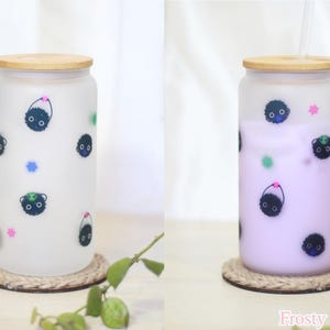 May include: Two frosted glass tumblers with bamboo lids and straws. The tumblers feature a black and white cartoon print of a creature with a small hat. One tumbler is filled with a light purple liquid, the other is empty. The text "Frosty Soots" is printed on the bottom right corner of the image.