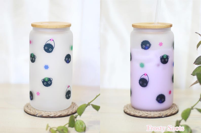 May include: Two frosted glass tumblers with bamboo lids and straws. The tumblers feature a black and white cartoon print of a creature with a small hat. One tumbler is filled with a light purple liquid, the other is empty. The text "Frosty Soots" is printed on the bottom right corner of the image.