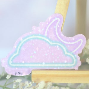 May include: A holographic sticker with a pink and blue cloud and crescent moon design. The sticker has a glittery finish and says "peel" at the bottom.
