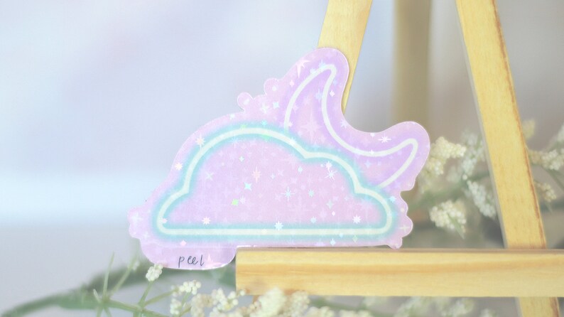 May include: A holographic sticker with a pink and blue cloud and crescent moon design. The sticker has a glittery finish and says "peel" at the bottom.
