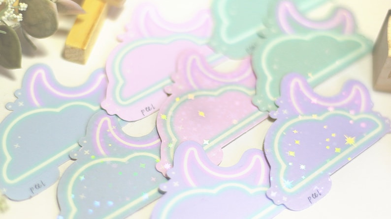 May include: A set of pastel-colored stickers with a moon and star design. The stickers are holographic and have a peel-off backing. The stickers say "peel" on the bottom.