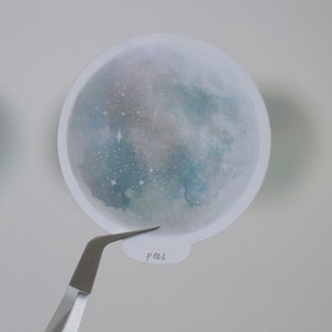 Puede incluir: Una pegatina redonda mate con un dise&ntilde;o de acuarela de una luna con estrellas. La pegatina es blanca con un dise&ntilde;o de acuarela azul y verde. El texto "pal" est&aacute; escrito en la pegatina.