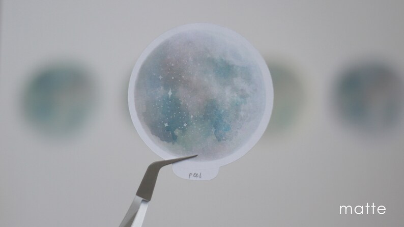 Puede incluir: Una pegatina redonda mate con un dise&ntilde;o de acuarela de una luna con estrellas. La pegatina es blanca con un dise&ntilde;o de acuarela azul y verde. El texto "pal" est&aacute; escrito en la pegatina.