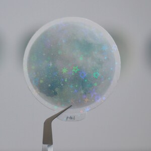 Puede incluir: Una pegatina redonda transparente con un acabado hologr&aacute;fico. La pegatina presenta un dise&ntilde;o de luna con estrellas y purpurina. La pegatina est&aacute; siendo sostenida por unas pinzas.