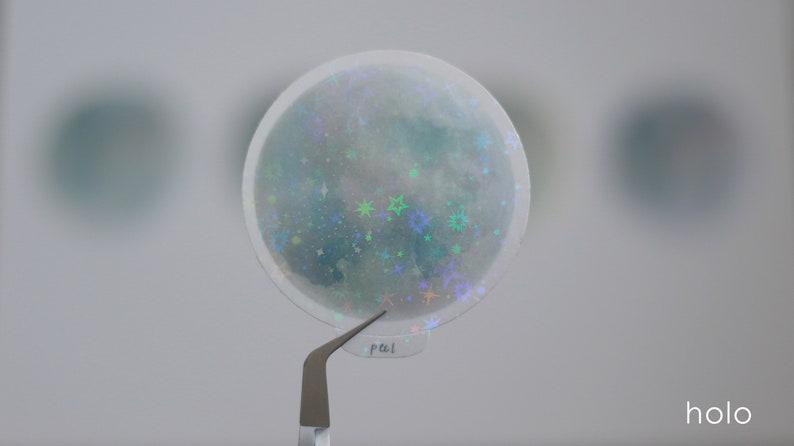 Puede incluir: Una pegatina redonda transparente con un acabado hologr&aacute;fico. La pegatina presenta un dise&ntilde;o de luna con estrellas y purpurina. La pegatina est&aacute; siendo sostenida por unas pinzas.