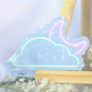 May include: A holographic sticker with a blue cloud and a pink crescent moon with stars. The sticker has the word "peel!" written on it.