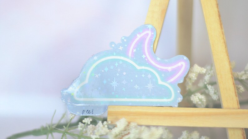 May include: A holographic sticker with a blue cloud and a pink crescent moon with stars. The sticker has the word "peel!" written on it.