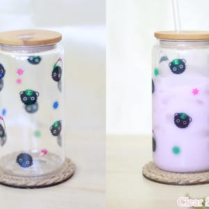 May include: Two clear glass tumblers with bamboo lids and black and green cartoon characters printed on them. One tumbler is empty and the other is filled with a light purple liquid. Both tumblers are sitting on brown rope coasters.