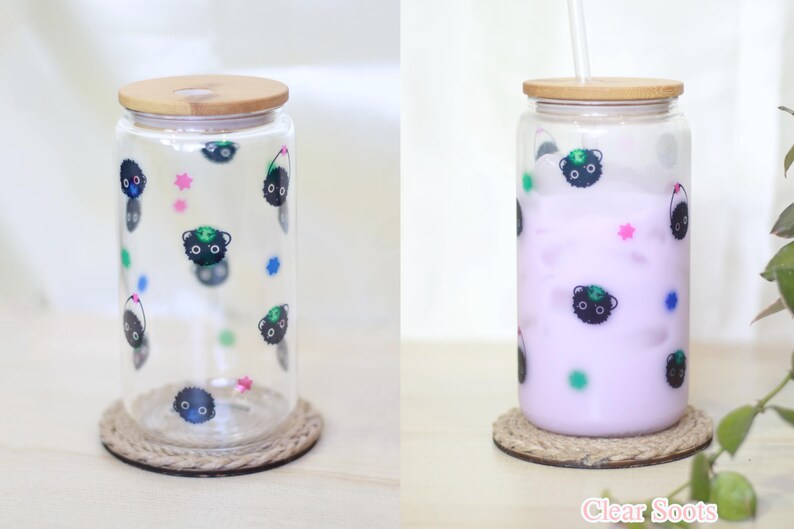 May include: Two clear glass tumblers with bamboo lids and black and green cartoon characters printed on them. One tumbler is empty and the other is filled with a light purple liquid. Both tumblers are sitting on brown rope coasters.