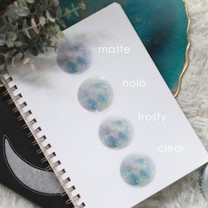 Puede incluir: Cuatro pegatinas en forma de luna con un dise&ntilde;o de galaxia, etiquetadas como "mate", "holo", "escarchado" y "transparente".