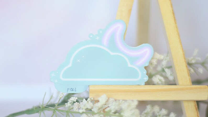 May include: A light blue cloud sticker with a white outline and a pink neon crescent moon. The sticker has a white "peel" label on the bottom.