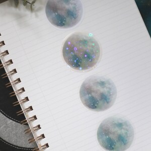 Puede incluir: Cuatro pegatinas redondas con un dise&ntilde;o de acuarela de una luna con estrellas. Las pegatinas est&aacute;n en una libreta blanca con una cubierta negra.