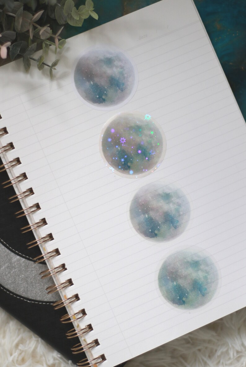 Puede incluir: Cuatro pegatinas redondas con un dise&ntilde;o de acuarela de una luna con estrellas. Las pegatinas est&aacute;n en una libreta blanca con una cubierta negra.