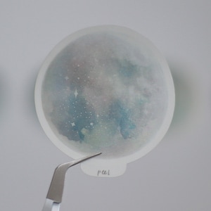Puede incluir: Una pegatina transparente con un dise&ntilde;o de acuarela de una galaxia azul y verde con estrellas blancas. La pegatina es redonda y tiene un borde blanco.