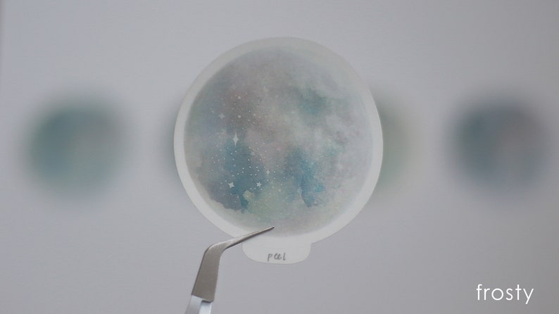 Puede incluir: Una pegatina transparente con un dise&ntilde;o de acuarela de una galaxia azul y verde con estrellas blancas. La pegatina es redonda y tiene un borde blanco.