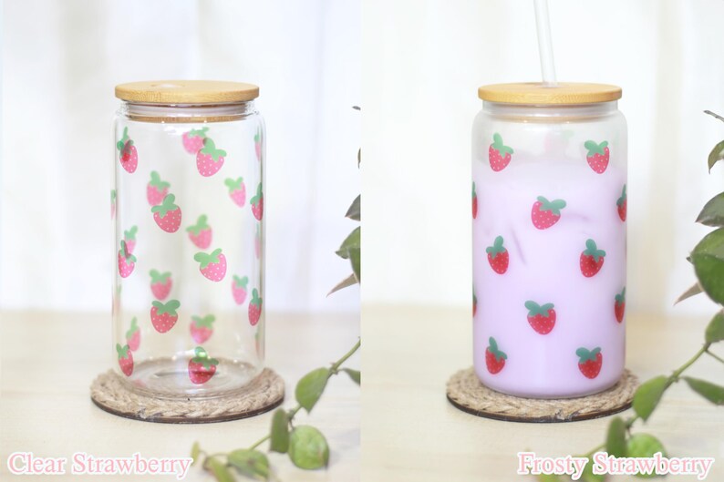 May include: Two glass jars with bamboo lids and strawberrry designs. The jar on the left is clear and the jar on the right is filled with a light purple liquid. Both jars are sitting on a brown rope coaster. The text "Clear Strawberry" is below the left jar and the text "Frosty Strawberry" is below the right jar.