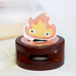 May include: A small, orange and pink fire spirit sticker with glitter, sitting on a wooden base. The sticker has two large eyes and the text "PUI" is visible on the bottom.
