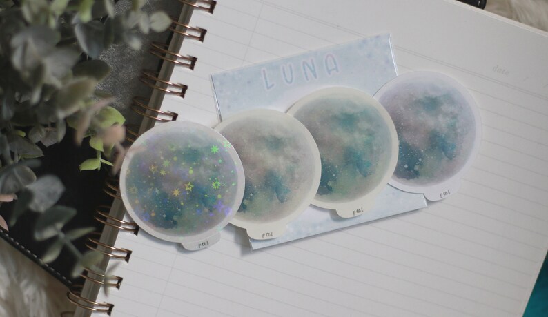 Puede incluir: Cuatro pegatinas de luna hologr&aacute;ficas iridiscentes con un dise&ntilde;o de acuarela azul y verde. Las pegatinas son redondas y tienen un borde blanco. El texto "LUNA" est&aacute; impreso en la parte superior de la hoja de pegatinas.