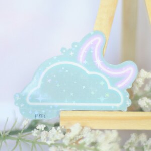 May include: A light blue cloud sticker with a pink neon crescent moon and glitter accents. The sticker is on a wooden easel with white flowers.