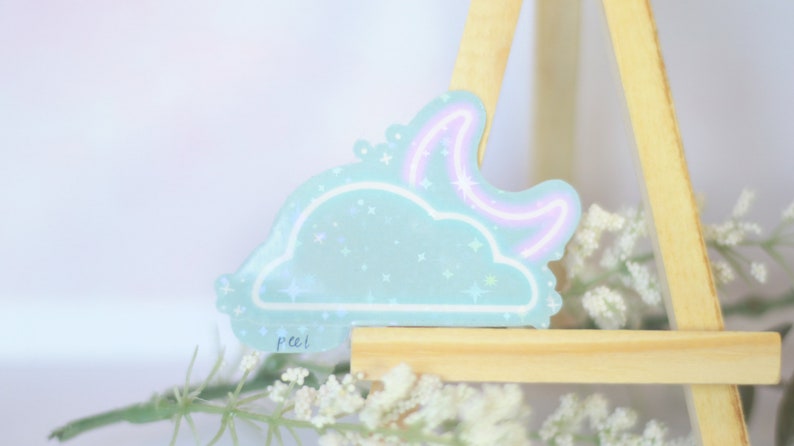 May include: A light blue cloud sticker with a pink neon crescent moon and glitter accents. The sticker is on a wooden easel with white flowers.
