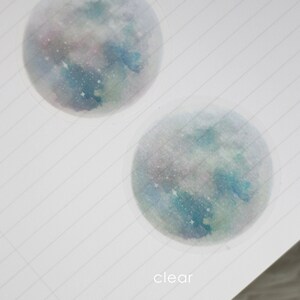 Puede incluir: Dos pegatinas redondas transparentes con un dise&ntilde;o de acuarela de una galaxia azul y verde con estrellas blancas. La pegatina superior est&aacute; etiquetada como "frosty" y la pegatina inferior est&aacute; etiquetada como "clear".