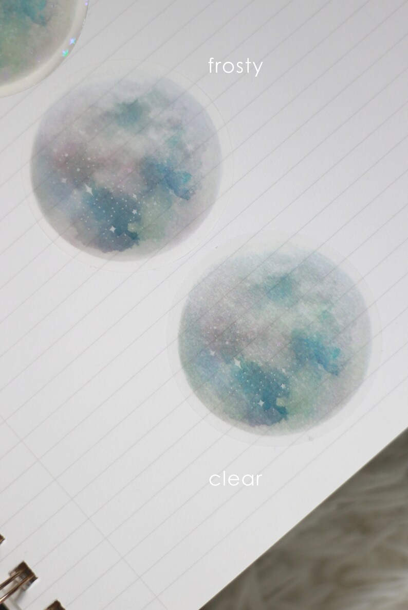 Puede incluir: Dos pegatinas redondas transparentes con un dise&ntilde;o de acuarela de una galaxia azul y verde con estrellas blancas. La pegatina superior est&aacute; etiquetada como "frosty" y la pegatina inferior est&aacute; etiquetada como "clear".
