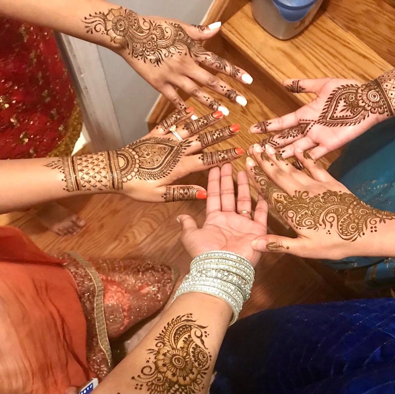 May include: Close-up of multiple hands adorned with intricate henna designs. The henna patterns feature floral and geometric motifs in shades of brown. Some nails are painted with bright colors, and silver bracelets are visible.