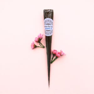 May include: A dark brown henna cone with a white label featuring the text "Henna By Lon Eucalyptus oil". The cone is surrounded by pink flowers on a light pink background. The henna cone is a traditional body art product.