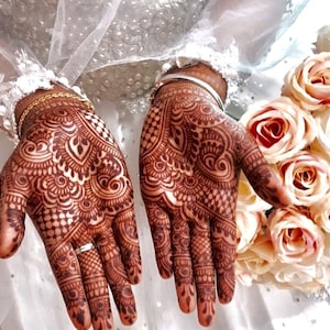 May include: Close-up of hands adorned with intricate henna designs in shades of brown. The detailed patterns cover the palms and fingers, with a wedding ring visible. A bouquet of light pink roses and a white veil are in the background.