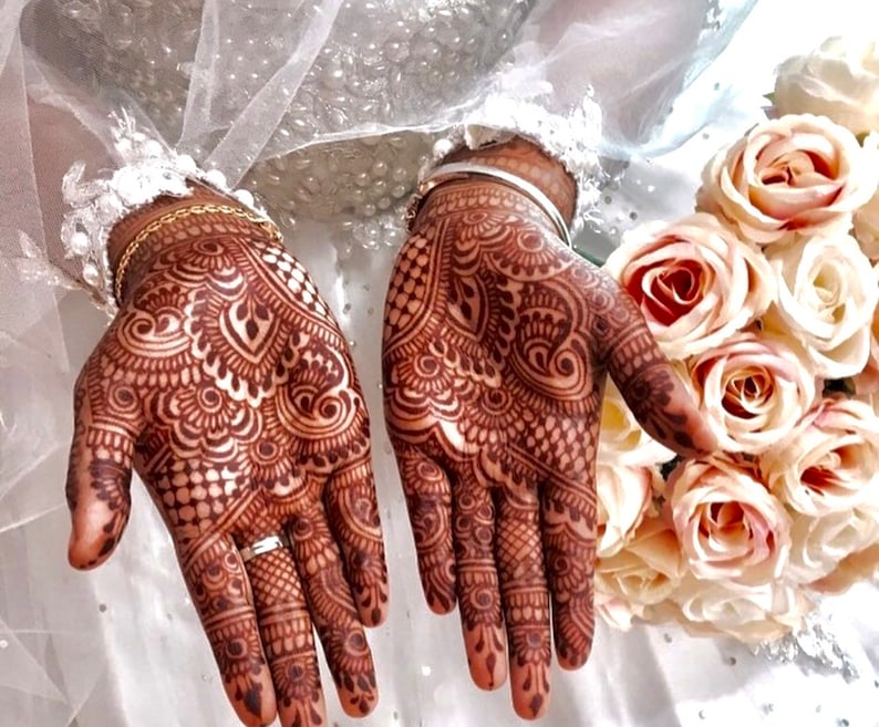 May include: Close-up of hands adorned with intricate henna designs in shades of brown. The detailed patterns cover the palms and fingers, with a wedding ring visible. A bouquet of light pink roses and a white veil are in the background.