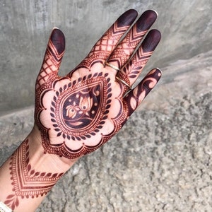 May include: Close-up of a hand adorned with intricate henna designs. The henna is a deep reddish-brown color, featuring floral and geometric patterns. The fingertips are stained a darker shade. Two gold rings are visible.