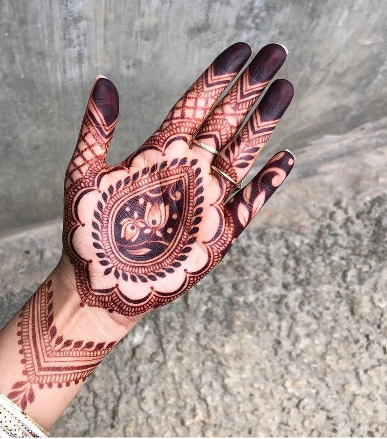 May include: Close-up of a hand adorned with intricate henna designs. The henna is a deep reddish-brown color, featuring floral and geometric patterns. The fingertips are stained a darker shade. Two gold rings are visible.