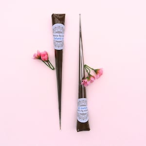 May include: Two henna cones with a floral arrangement on a pink background. The henna cones are dark brown with a white and blue label that reads "Henna By Lo Eucalyptus oil". The cones are next to a metal applicator.